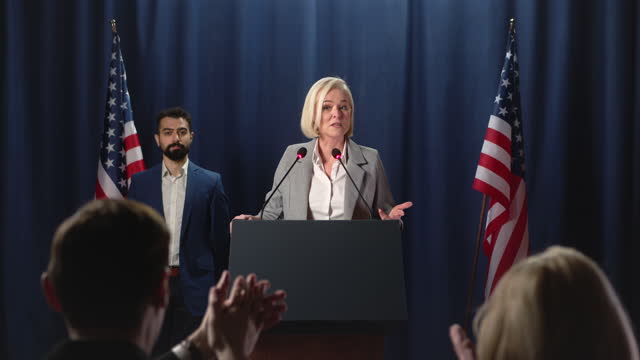A blond female politician ascends  the scene and starts a speech