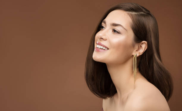 joyeux souriant modèle profil vue latérale. femme brune beauté avec maquillage nu peau lisse et boucle d’oreille dorée sur fond brun - health or beauty photos photos et images de collection