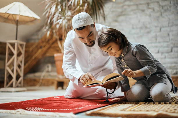 Happy Muslim girl and her father reading Koran at home. Middle Eastern man teaching his daughter to read from Koran while praying at home. father allah stock pictures, royalty-free photos & images