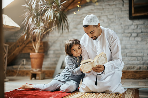 Muslim little girl sitting next to her father who is reading her Quran at home.