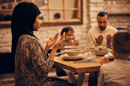 African American Muslim woman praying with her family at dining table on Ramadan.