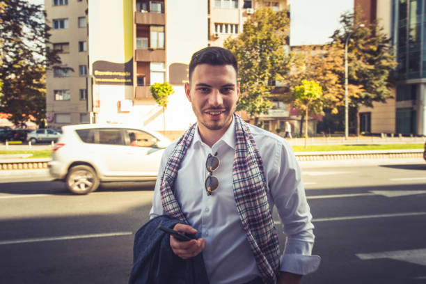 A good looking businessman is standing on the street, he is looking front and smiling while holding his coat and phone in hands