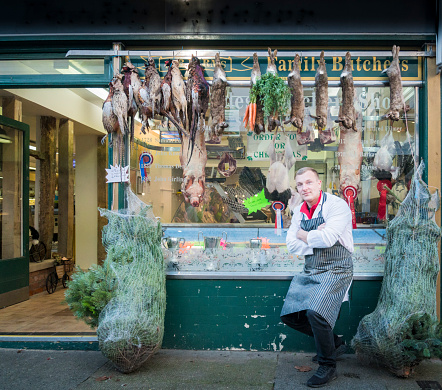 A male butcher standing outside his shop window with pheasants and rabbits hanging up for display