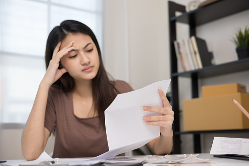 A young Asian woman is stressed because she looks at many expense bills and she has no money to pay them. Hand on head feeling very serious depressed.