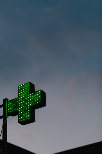 A green lit pharmacy cross against dark cloudy in the evening. Silhouette of a building on the bottom of a frame. Low angle shot