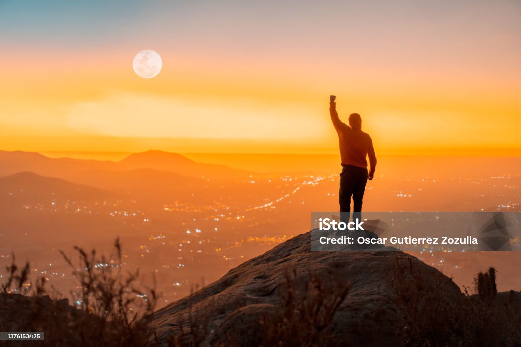 person standing on the top of the mountain with hand up, back view, over the city at sunset Success Stock Photo