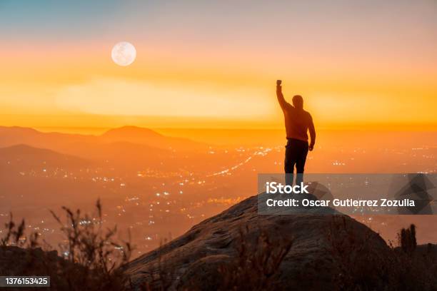 Person Standing On The Top Of The Mountain With Hand Up Back View Over The City At Sunset Stock Photo - Download Image Now