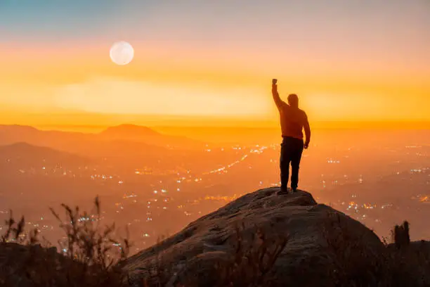 Photo of person standing on the top of the mountain with hand up, back view, over the city at sunset