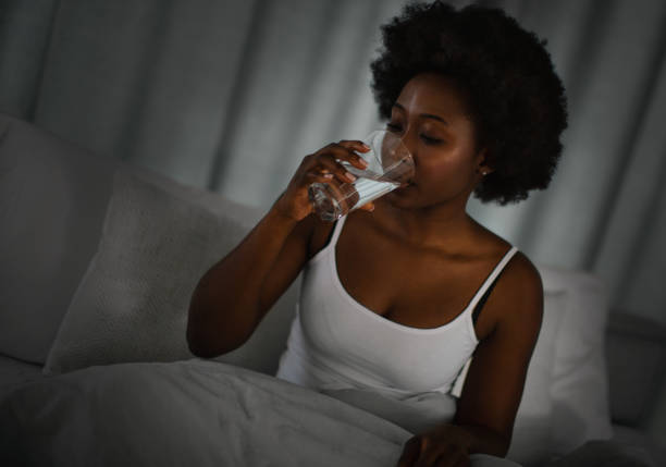 Young woman getting up during the night because of thirst. She is sitting on the bed and drinking water.