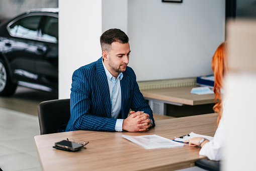 Handsome young businessman talking with sales person at car showroom while buying his new or used vehicle and signing buyer's contract.