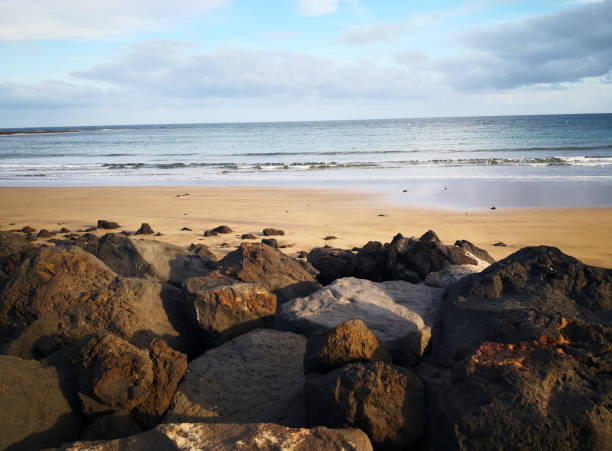 Idyllic holiday landscape for relaxation rocas de lava de textura negra en los rayos del sol de la tarde en el borde de la playa de arena en lanzarote, islas canarias. las piedras negras y marrones oscuras contrastan con la arena amarilla y el agua azul del océano atlántico y el cielo con nubes
