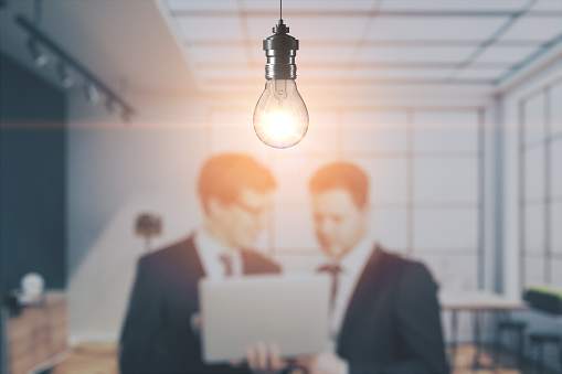 Blurry businessmen with laptop working together on abstract office interior background with glowing light bulb. Work ideas and innovations concept. Mock up place for your advertisement