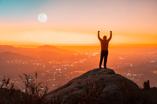 person silhouette on the top of the mountain with hands raised, back viev, sunset background