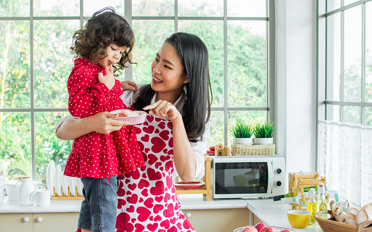 Beautiful Asian woman wearing apron and little cute daughter girl, smiling with happiness, having breakfast, playing together in kitchen at home on Valentine day in morning. Lifestyle, family concept