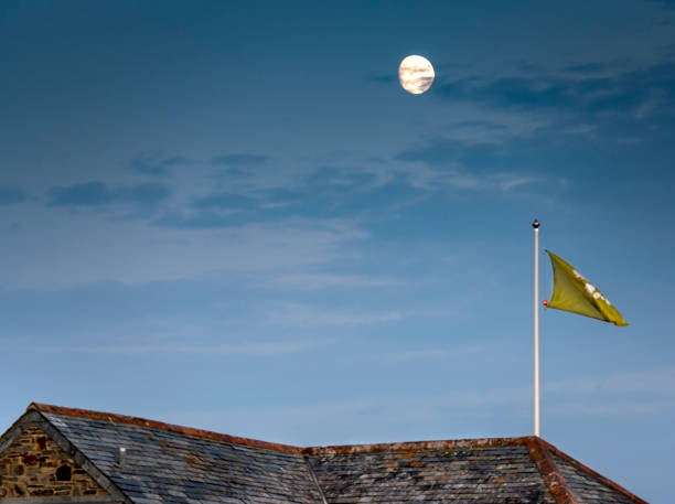 eine national trust-flagge weht hoch auf einem kornischen dach, mit dem silbernen mond dahinter, vor einem sich verdunkelnden himmel. - cornish flag stock-fotos und bilder