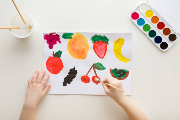 Baby hand holding paint brush and drawing different colorful fruit and berry shapes on white paper on light table background. Closeup. Point of view shot. Toddler development. Top down view. stock photo