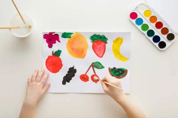 Baby hand holding paint brush and drawing different colorful fruit and berry shapes on white paper on light table background. Closeup. Point of view shot. Toddler development. Top down view.