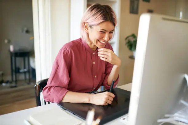 Photo of Indoor shot of cheerful happy young female with stylish pinkish hair laughing while working from home, sitting at desk with computer and graphic tablet, retouching images or drawing animation