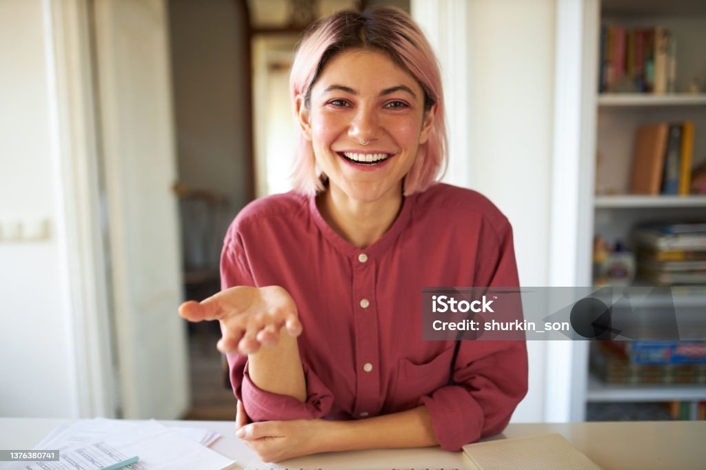 Happy cute teenage girl with pinkish hair smiling broadly while talking to friend online using video conference service. Stylish young female influencer recording vlog via webcam on electronic device Talking Stock Photo