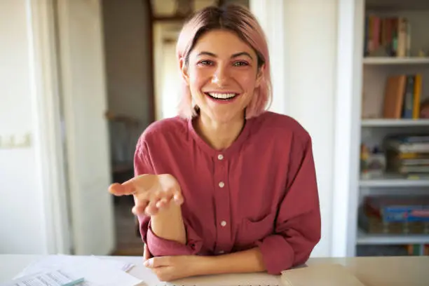 Photo of Happy cute teenage girl with pinkish hair smiling broadly while talking to friend online using video conference service. Stylish young female influencer recording vlog via webcam on electronic device