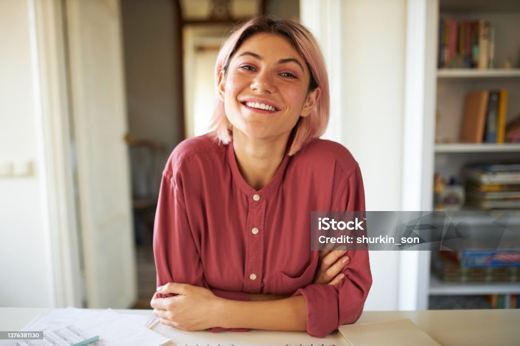 Positive cheerful young woman posing in cozy home interior, sitting at table with papers, working distantly, looking at camera with broad smile, having online group meeting with colleagues Interview - Event Stock Photo