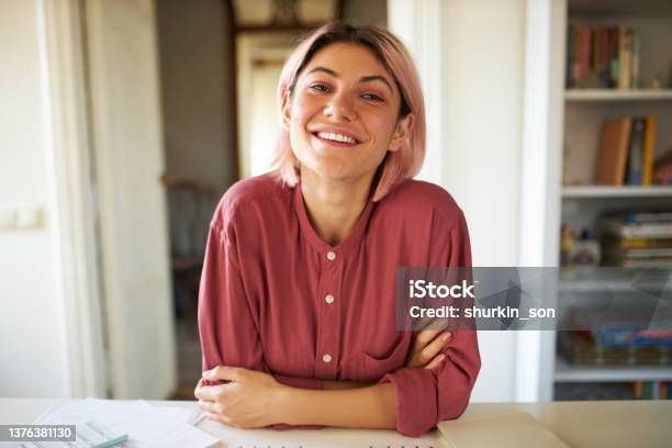 Positive Cheerful Young Woman Posing In Cozy Home Interior Sitting At Table With Papers Working Distantly Looking At Camera With Broad Smile Having Online Group Meeting With Colleagues Stock Photo - Download Image Now