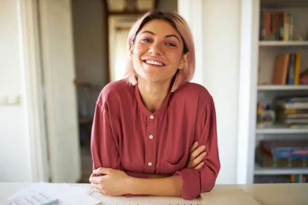 Photo of Positive cheerful young woman posing in cozy home interior, sitting at table with papers, working distantly, looking at camera with broad smile, having online group meeting with colleagues