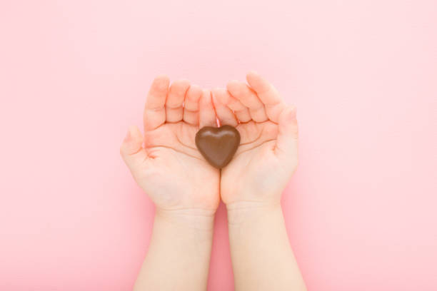 herzform von schwarzen dunklen schokoladenbonbons in baby mädchen geöffnet handflächen auf hellrosa tischhintergrund. pastellfarben. süßer snack. closeup. standpunkt aufgenommen. ansicht von oben nach unten. - heart shape child human hand childhood stock-fotos und bilder