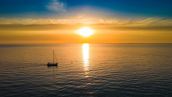 Yacht sails to the horizon - photograph of a bright sunset with a yacht sailing on the sea. Beautiful sunset at sea over the horizon with clouds in the sky. A small boat sails into the sunset. A sunset over the ocean. Reflection of a sunset at sea. Sunrise at sea. Sunset in the clouds over the sea horizon. Little white boat floating on the water towards the horizon in the rays of the setting sun. Beautiful clouds with yellow highlights.