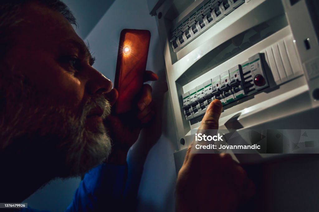 Energy crisis. Man in complete darkness holding a phone to investigate a home fuse box during a power outage. Blackout concept. Blackout Stock Photo