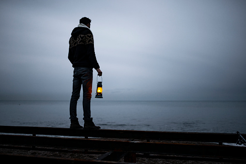 young man standing on the beach in cold weather, holding lighthouse, looking at horizon.