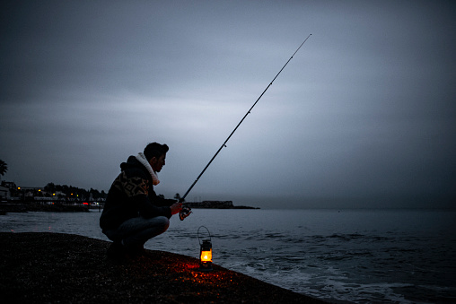 Teenager crouching fishing with a sailor's lighthouse in cloudy weather at sunset on the beach.