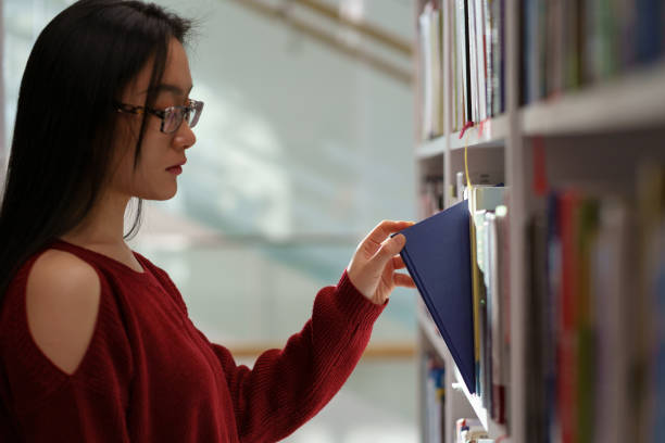 istock Asian student girl picking up book from shelf while standing between library bookcases 1376510532
