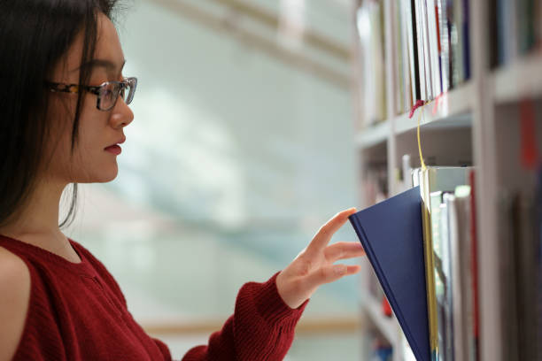 istock Asian student girl picking up book from shelf while standing between library bookcases 1376510546