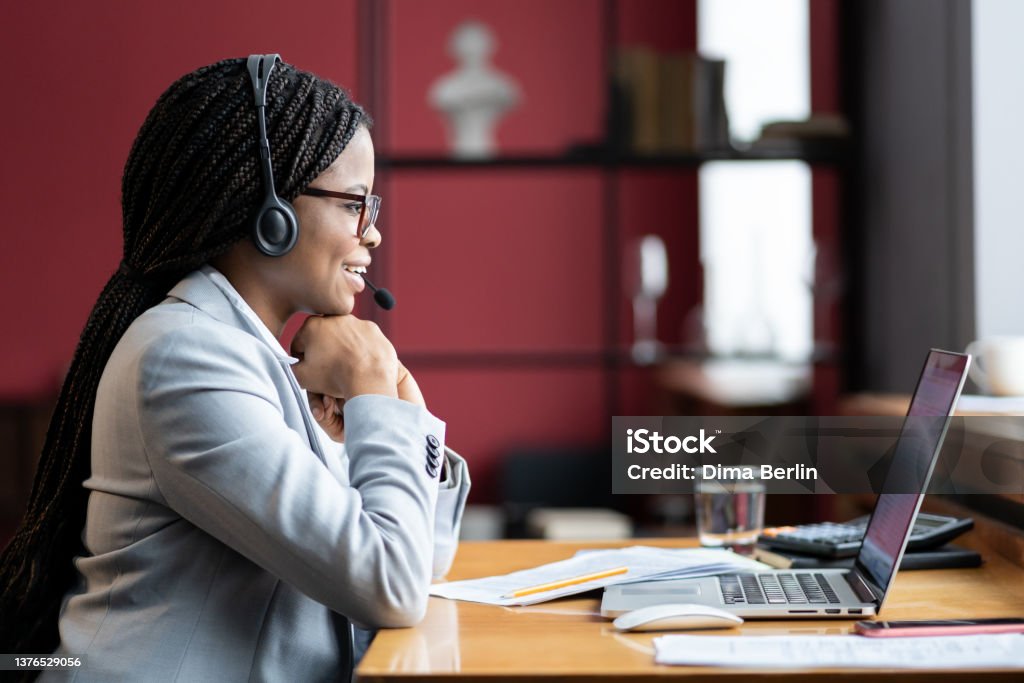 Young smiling african woman mother on maternity leave in wireless headset working remotely from home Young positive black woman mother on maternity leave in headset working remotely on laptop, pleasant remote female employee discussing project with collegues durig virtual meeting. Selective focus Using Phone Stock Photo
