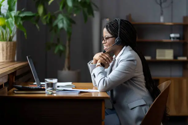 Young friendly black woman remote call center worker in headset with microphone broadly smiling while communicating with customers, looking at laptop screen while consulting clients remotely from home