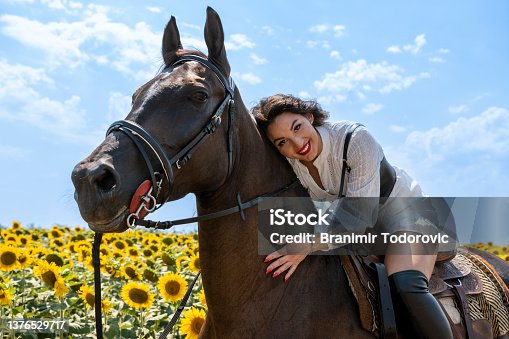 istock Cheerful brunette horseback riding. She is wearing traditional white dress and long black boots. Rural scene. Sunny day. Sunflower field in background. 1376529717
