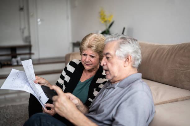 senior couple paying bills using smartphone at home - common serious couple men imagens e fotografias de stock