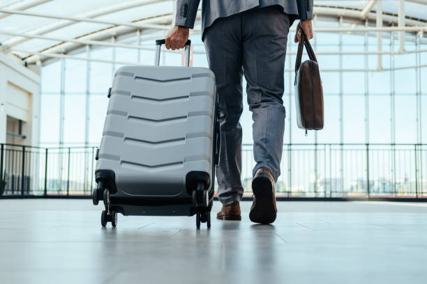 An Anonymous Business Man Walking With Luggage in the Airport Low angle view of unrecognizable businessman in gray suit walking through airport terminal with briefcase and suitcase. business travel stock pictures, royalty-free photos & images