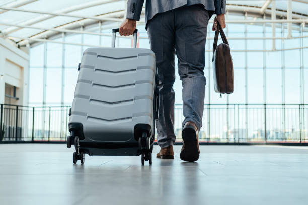 An Anonymous Business Man Walking With Luggage in the Airport Low angle view of unrecognizable businessman in gray suit walking through airport terminal with briefcase and suitcase. hand luggage stock pictures, royalty-free photos & images