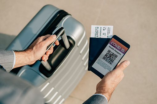 Traveling During Coronavirus Pandemic: High angle view of unrecognizable businessman hands holding his luggage, passport and phone with vaccine immunization certificate QR code.