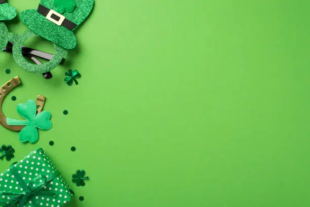 Top view photo of st patricks day decorations hat shaped party glasses horseshoe with green shamrock clover shaped confetti and giftbox on isolated pastel green background with copyspace