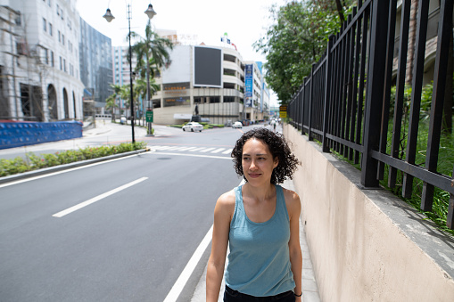 A young mixed race woman takes a walk around the block while in isolation during the Covid-19 pandemic lockdown. The usually busy street and urban neighborhood is empty. The smiling woman is savoring this peaceful moment outdoors. She is social distancing and is not wearing a face mask.