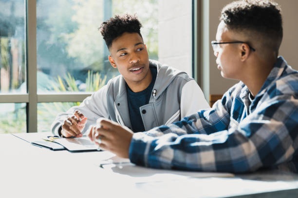 Two male teenage friends talk together during class Sitting at the same table, the two male teenage friends talk during class. young black boys stock pictures, royalty-free photos & images