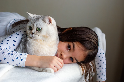 A little girl lies sad with a white cat on the edge of the bed, hugs him. Copy space