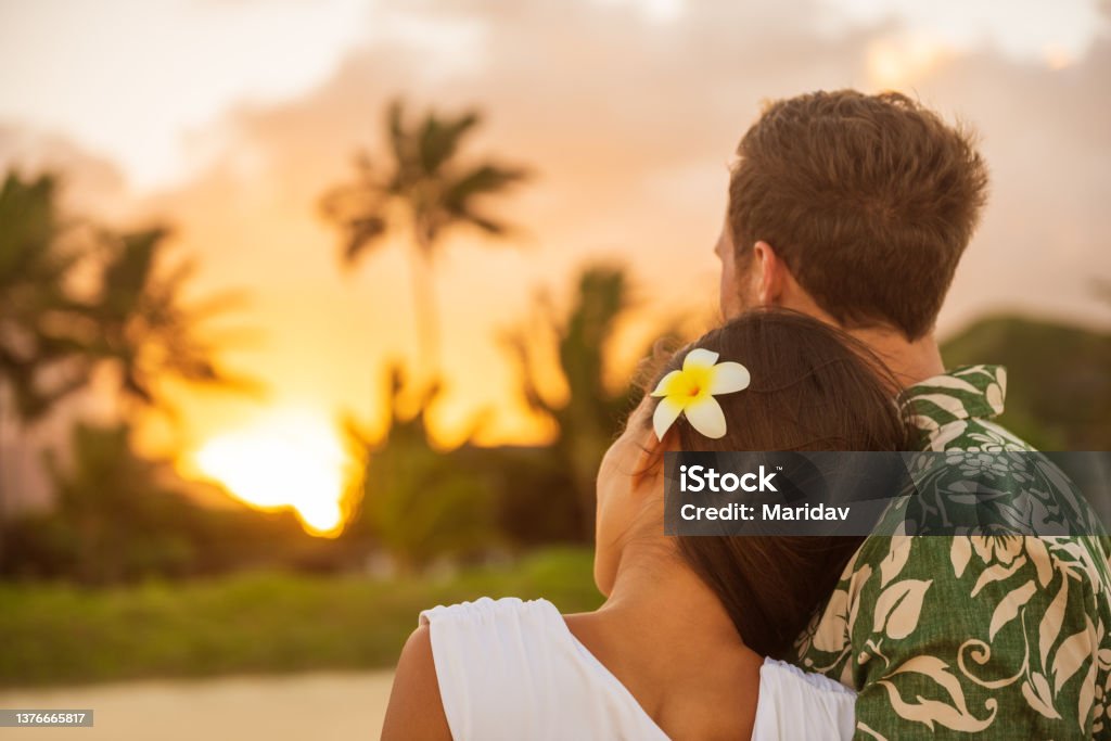 Romantic couple relaxing watching sunset on beach stroll view from back. Woman resting head on lover's shoulder on honeymoon vacation travel in summer Hawaii destination. Newlyweds people. Hawaii Islands Stock Photo