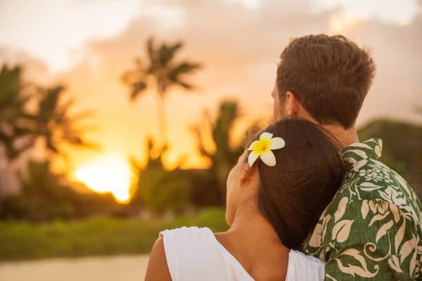 Romantic couple relaxing watching sunset on beach stroll view from back. Woman resting head on lover's shoulder on honeymoon vacation travel in summer Hawaii destination. Newlyweds people. stock photo