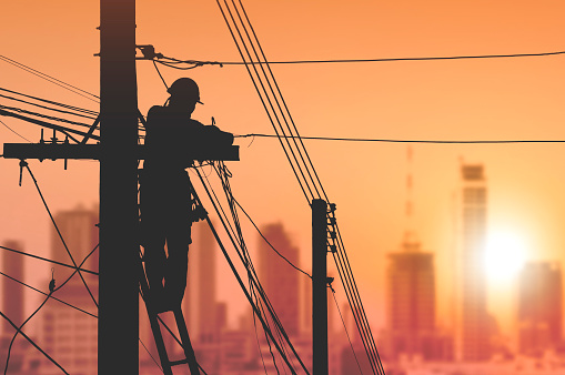 Silhouette electrician on ladder is installing cable lines to connecting internet signal on electric power pole with blurred cityscape view in sunrise sky background