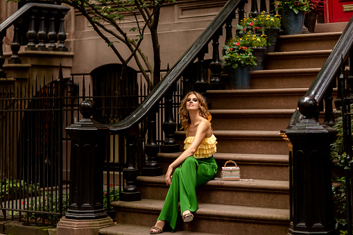 Slim red-haired caucasian young woman in stylish bright yellow top and green trousers elegant sandals sitting on the potted flowers porch stairs on Manhattan West village.