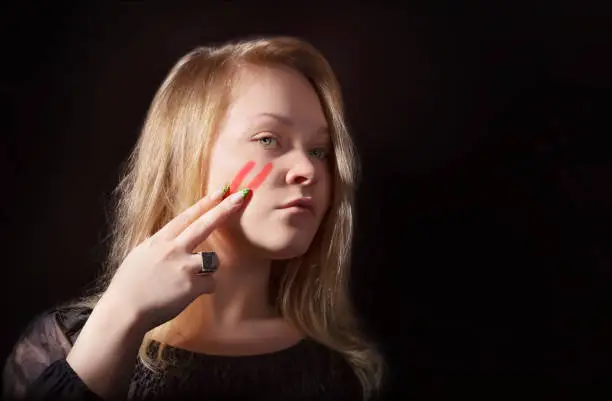 A young girl draws two stripes on her cheek with her fingers using lipstick War paint Black background